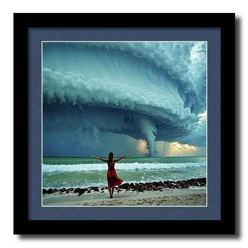 Digital artwork framed print of a woman in a red dress stands on a beach with her arms outstretched, facing an approaching storm with massive, swirling clouds. 